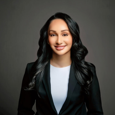 Professional headshot of Priyanka Shah, a woman with long black hair styled in loose curls, wearing a black blazer over a white top. She is smiling warmly and posed against a dark gray background.