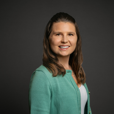Professional headshot of Rachel Inman, a woman with long wavy brown hair, wearing a mint green cardigan over a white top. She is smiling and posed against a dark gray background.
