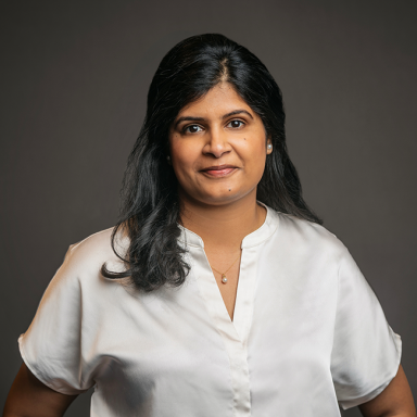 Professional headshot of Sruthi Meka, a woman with long black hair, wearing a silky white blouse and a delicate pendant necklace. She has a neutral expression and is posed against a dark gray background.