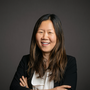Professional headshot of Sung Hee Choe, a woman with long brown hair, wearing a white ruffled top under a black blazer. She is smiling with arms crossed and posed against a dark gray background.