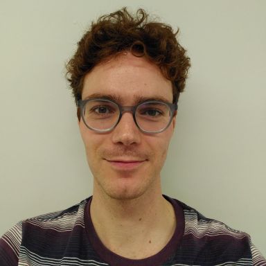 A person with short curly brown hair, wearing gray-framed glasses and a striped shirt, smiling slightly in front of a plain light-colored background.