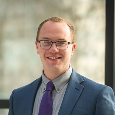 Professional headshot of smiling man in blue suit with purple tie and glasses