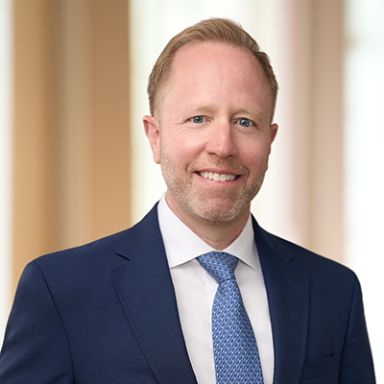 Professional headshot of smiling man in navy suit with blue patterned tie