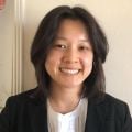 A smiling female of Asian heritage with short black hair wearing a black suit jacket over a white shirt against a cream background