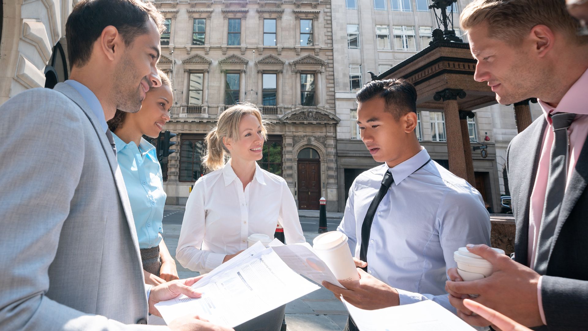 ChatGPT said:  A diverse group of professionals gathered outdoors in a city setting, engaging in a collaborative discussion while reviewing documents. The group includes both men and women dressed in business attire, holding coffee cups. The background features classic architecture and city streets. The scene reflects teamwork, professional growth, and community engagement, fitting for an economic mobility alliance.