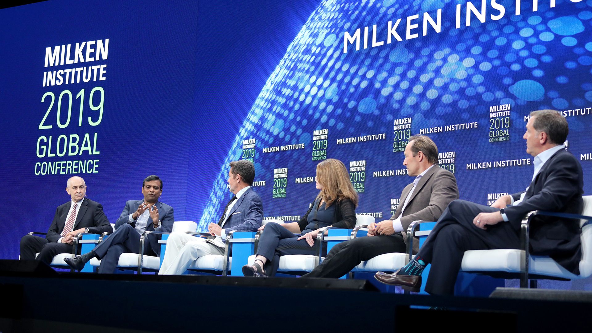 A panel of six speakers sits on stage at the Milken Institute 2019 Global Conference, with a large digital backdrop displaying the conference name and logo.