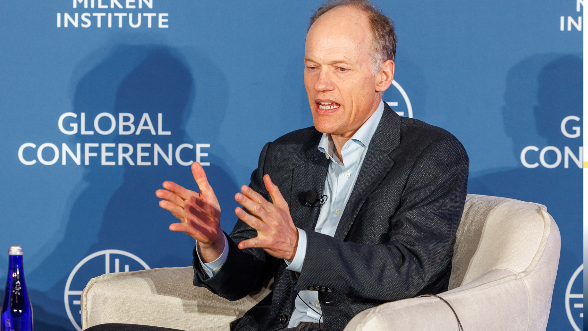 Sebastian Mallaby in suit gesturing while speaking at Global Conference panel, seated in white chair