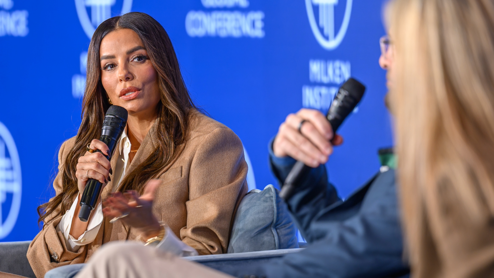 Woman in beige blazer speaks at press conference with blue branded backdrop