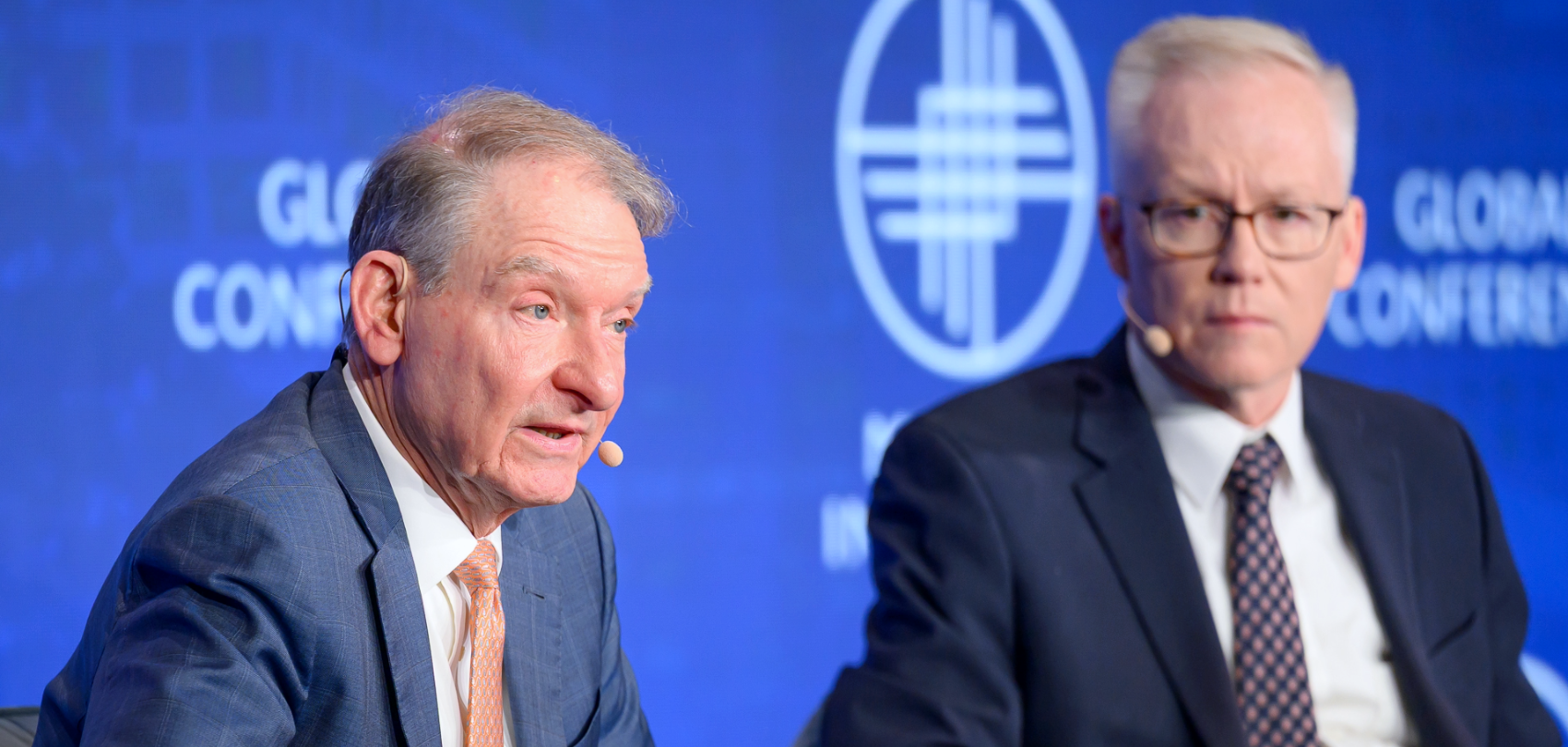 Two men in suits at a NATO conference, one speaking and one listening, against a blue background with NATO logo