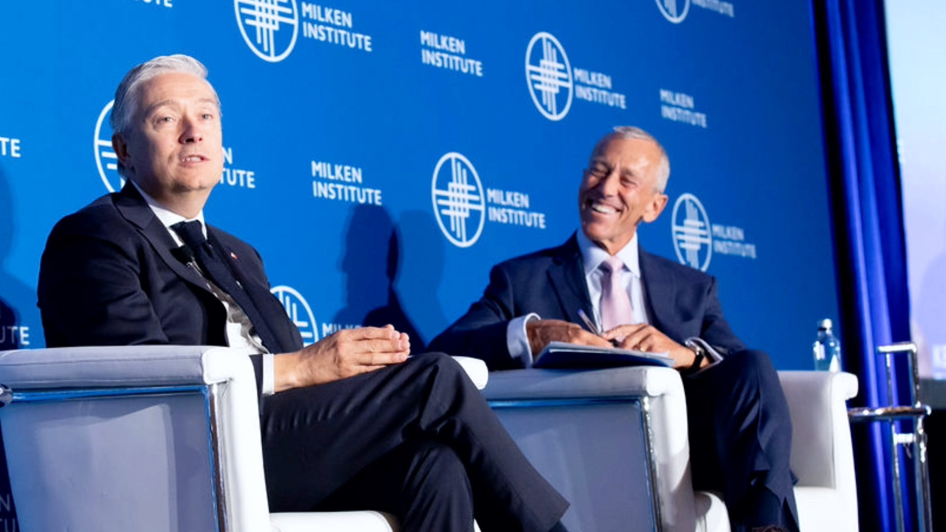 Two professionals in business attire seated in white chairs having a discussion at a corporate event with blue backdrop