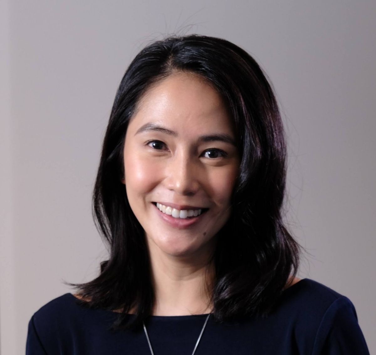 Smiling medium-skinned, female-presenting individual with medium length dark hair wearing a black shirt on a grey backdrop.