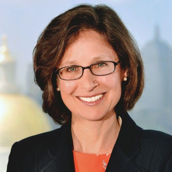 A smiling female-presenting person wearing eye glasses and a black blazer against a blurred background.