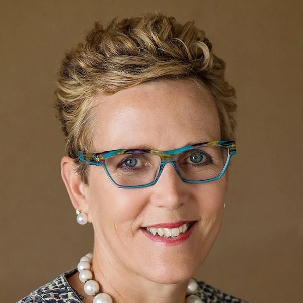 A smiling light-skinned female-presenting person with short, light hair wearing a pearl necklace against a plain brown background