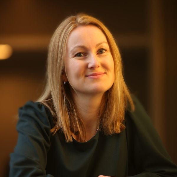 Smiling light-skinned female-presenting person with light hair wearing a black shirt against a blurred background