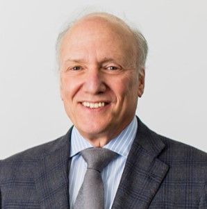 A smiling Caucasian male-presenting person with gray hair wearing a dark gray suit jacket and tie against a light gray background