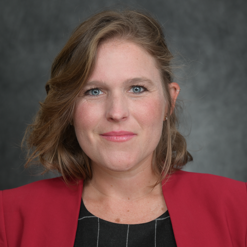 Light-skinned female-presenting person with light hair in a red jacket and black checkered shirt with gray gradient background