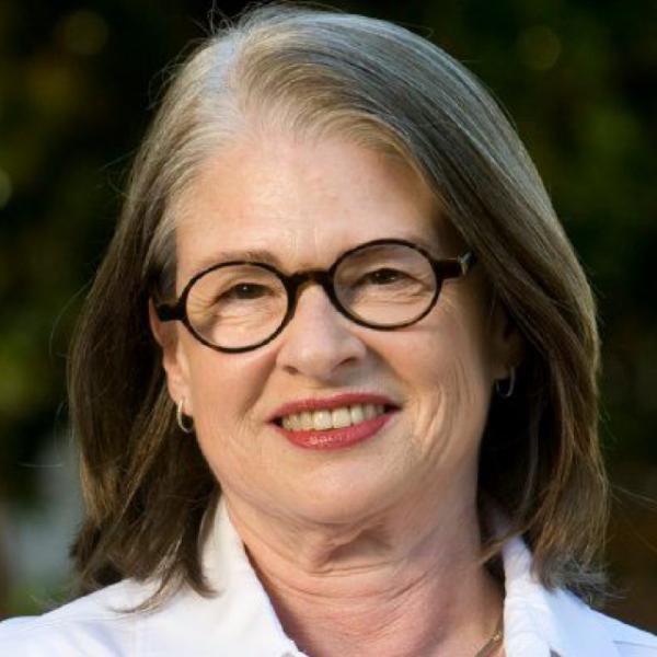 A female-presenting person with shoulder-length hair wearing a white top against a blurred background.