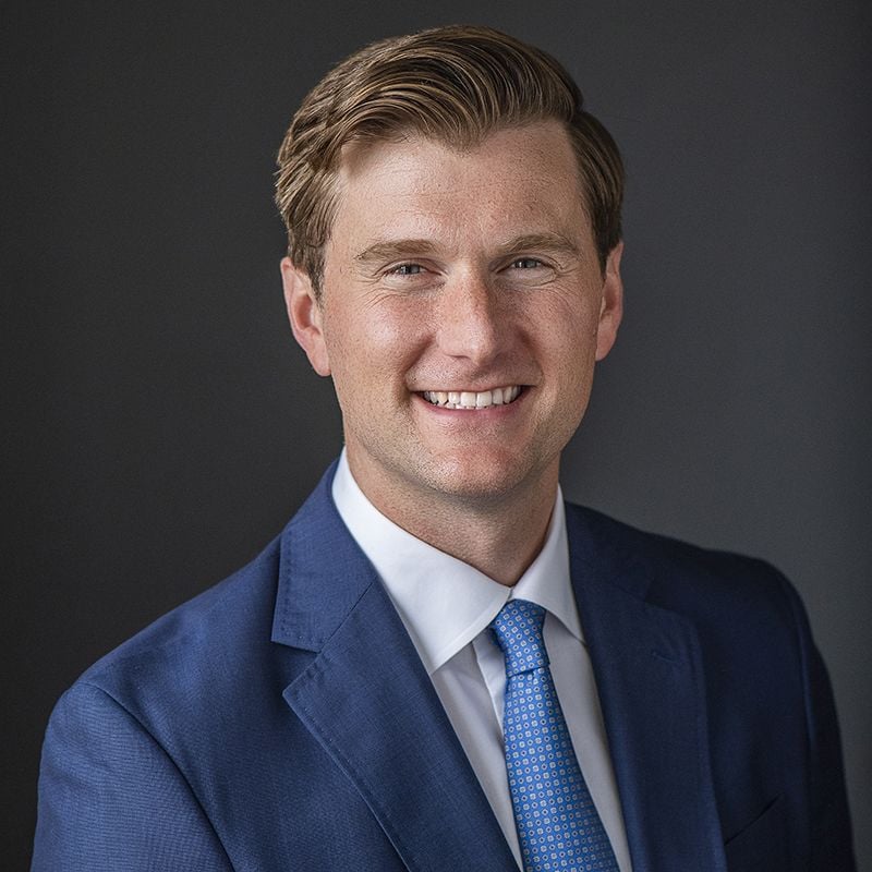 Smiling light-skinned male-presenting person wearing a blue suit with blue tie and white button-up against a dark gray background