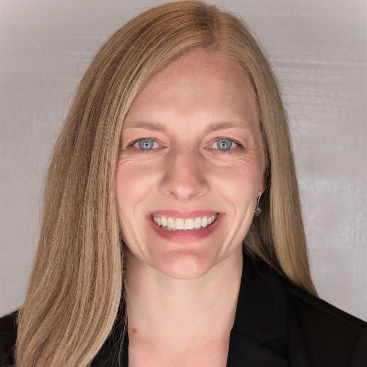 A smiling light-skinned female-presenting person with light hair wearing a black jacket against a light gray background