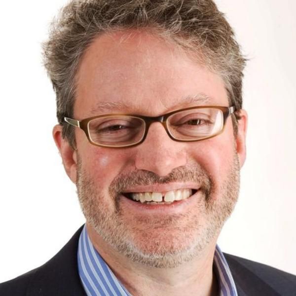 A smiling male-presenting person wearing eye glasses and a black blazer against a white background.