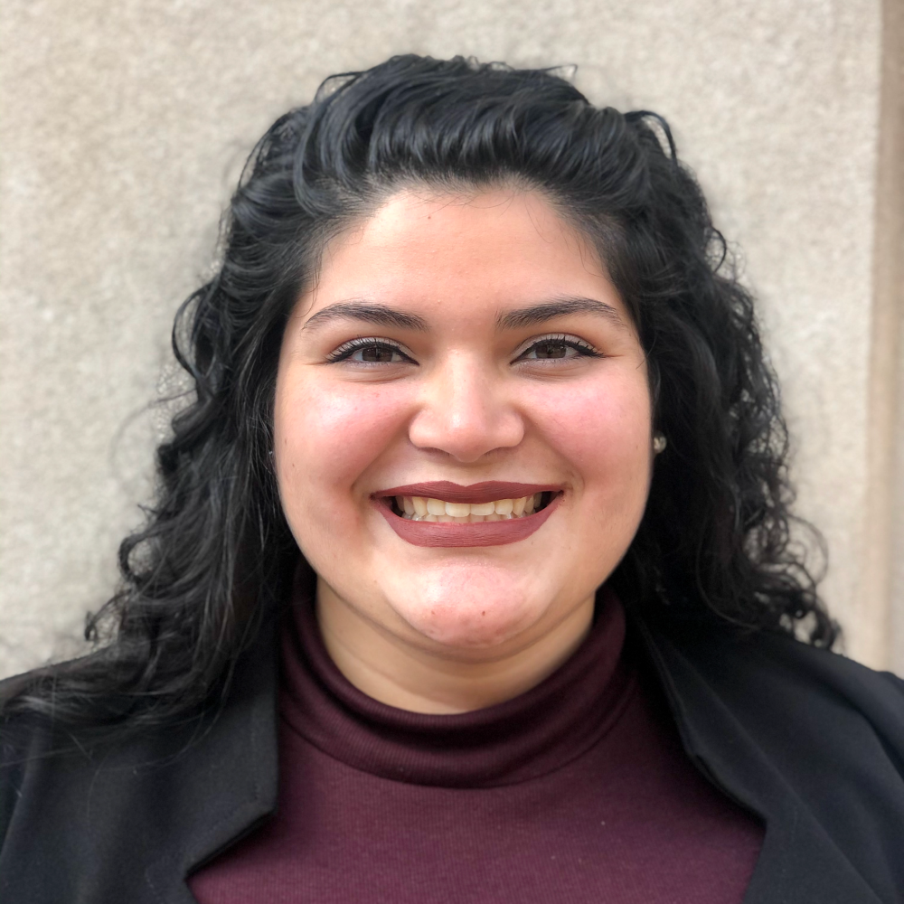 A smiling female-presenting individual person with black hair wearing a dark suit jacket over a maroon shirt with a light brown background