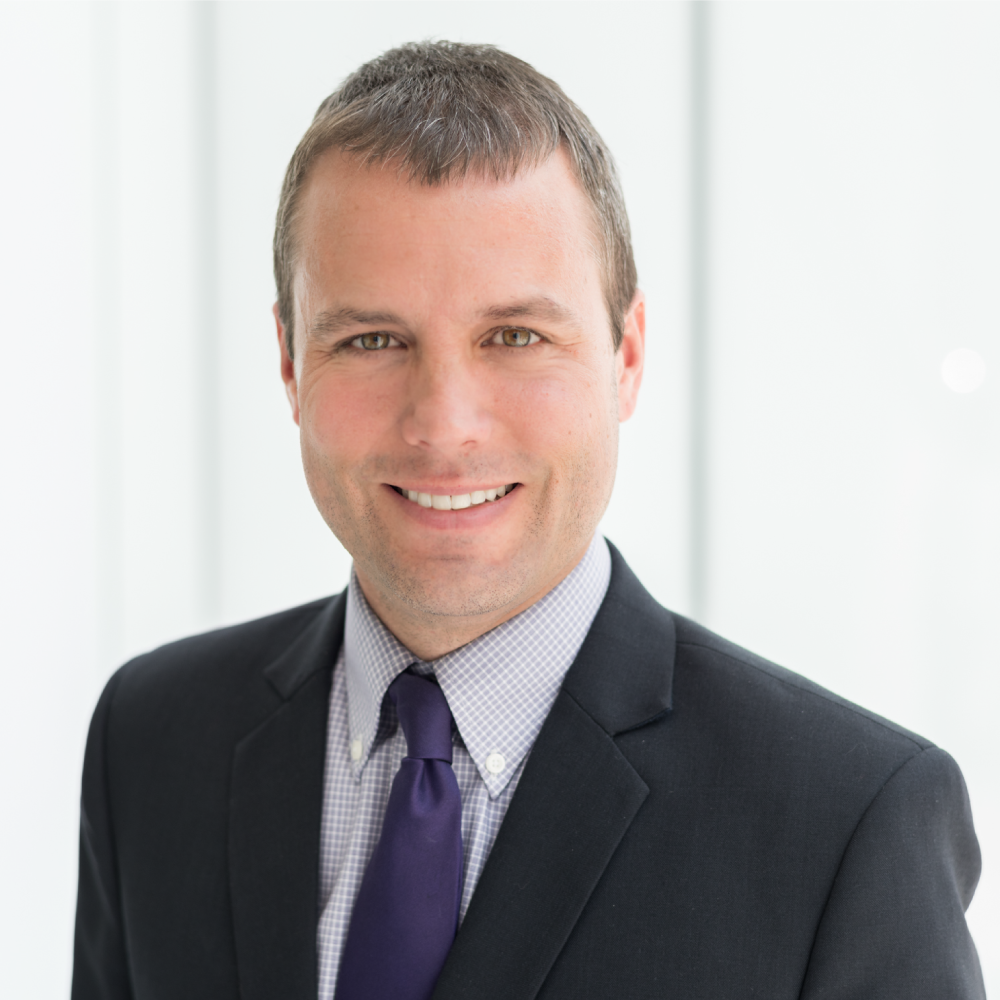 Smiling, male-presenting individual with a dark suit and tie against a white background