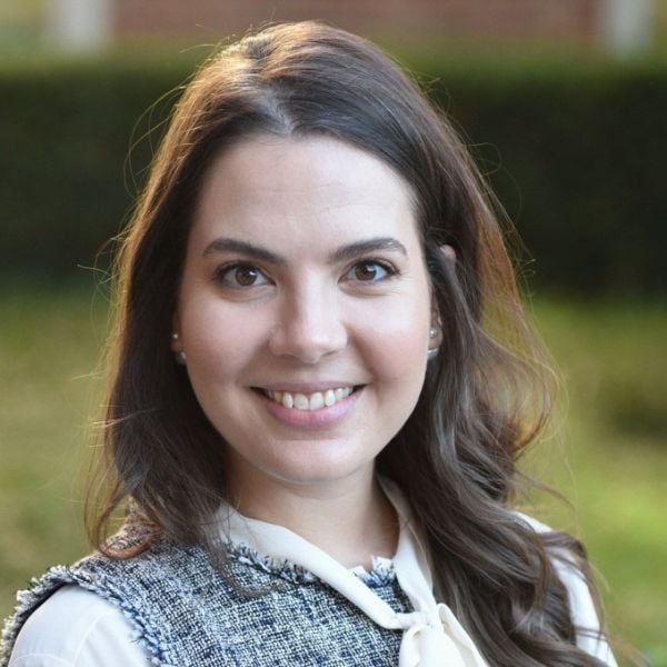 A smiling light-skinned female-presenting person with brown hair in a gray and white shirt against an outdoor backdrop.