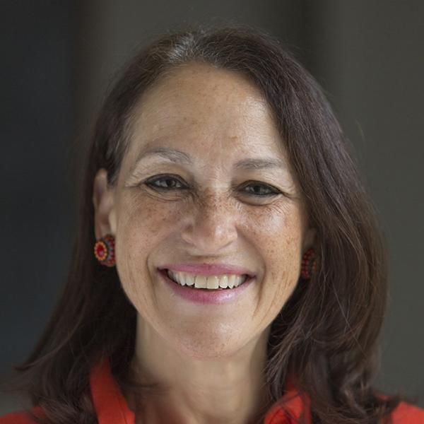 A smiling light-skinned female-presenting person with long, dark hair wearing a red shirt against a dark, blurred background