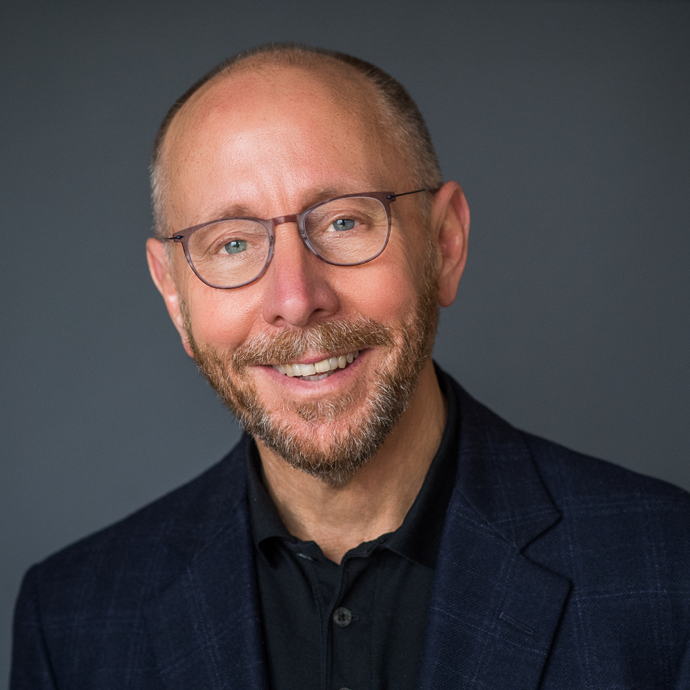 A smiling light-skinned, male-presenting person wearing a dark suit jacket over a dark shirt and glasses against a dark gray background