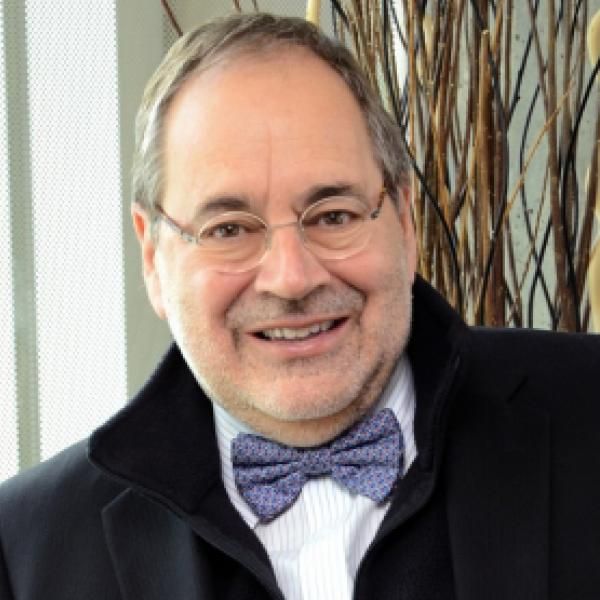 A smiling male-presenting person wearing eye glasses and a black blazer with a bow tie against a light background.