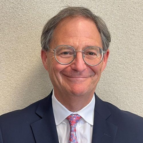 A smiling light-skinned male-presenting person with short, gray hair wearing a suit against a light background