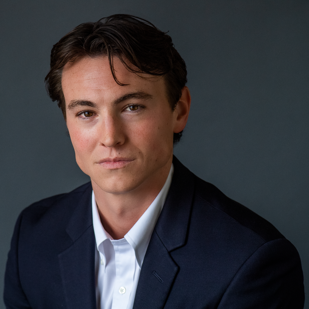 A male-presenting person with dark brown hair, wearing a dark suit and white button-up against a gray background