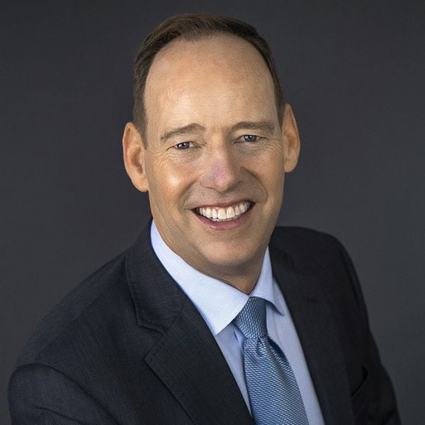 Smiling light-skinned, male-presenting individual with short dark hair wearing a dark suit on a grey backdrop.