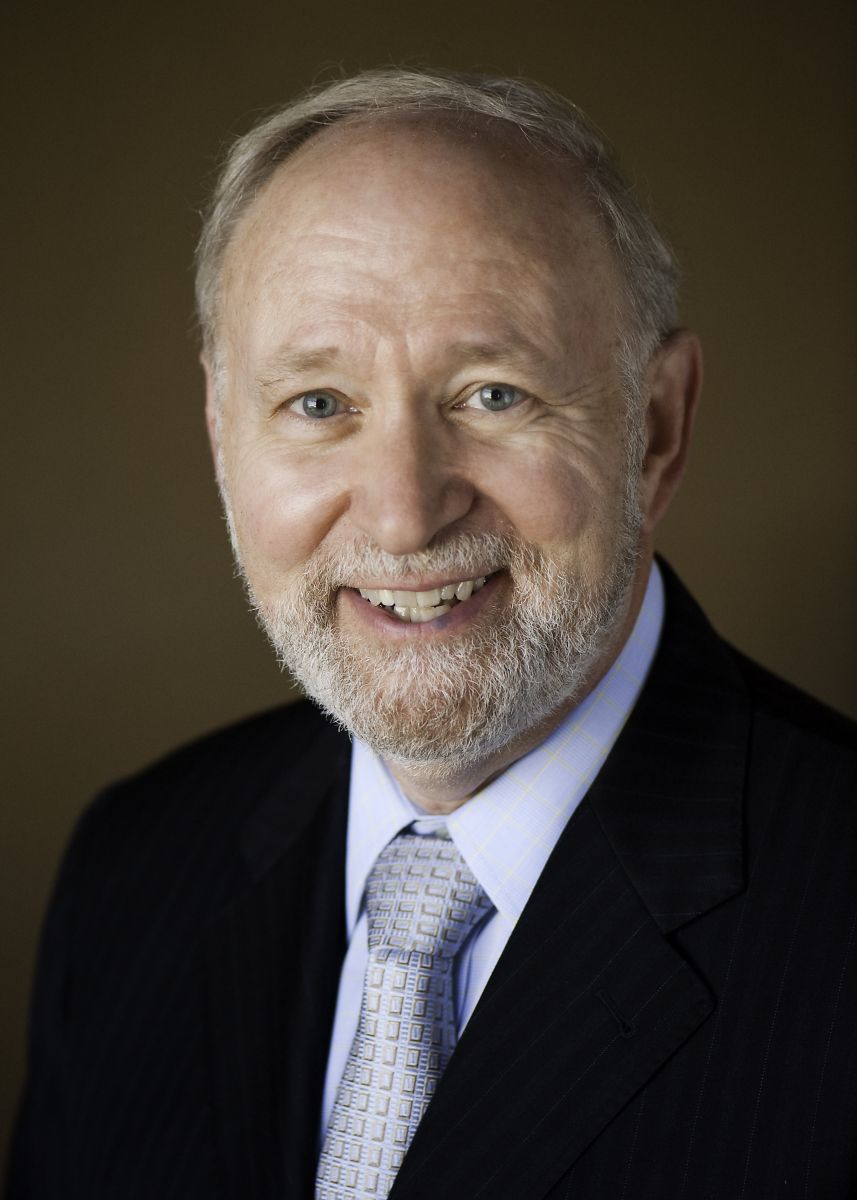 Smiling light-skinned, male-presenting individual with short grey hair wearing a dark suit on a brown backdrop.