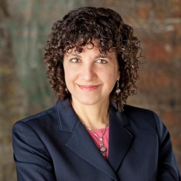 A smiling female-presenting person with curly hair wearing a dark blazer against a blurred background.