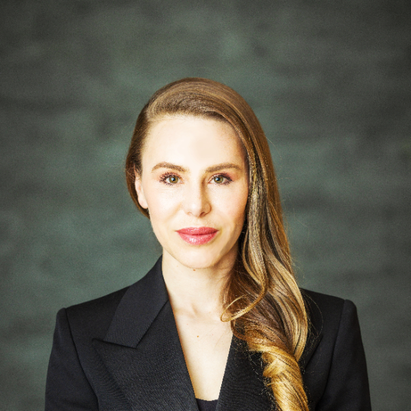 A light-skinned female-presenting person with long, red hair wearing a suit against a blurred dark background