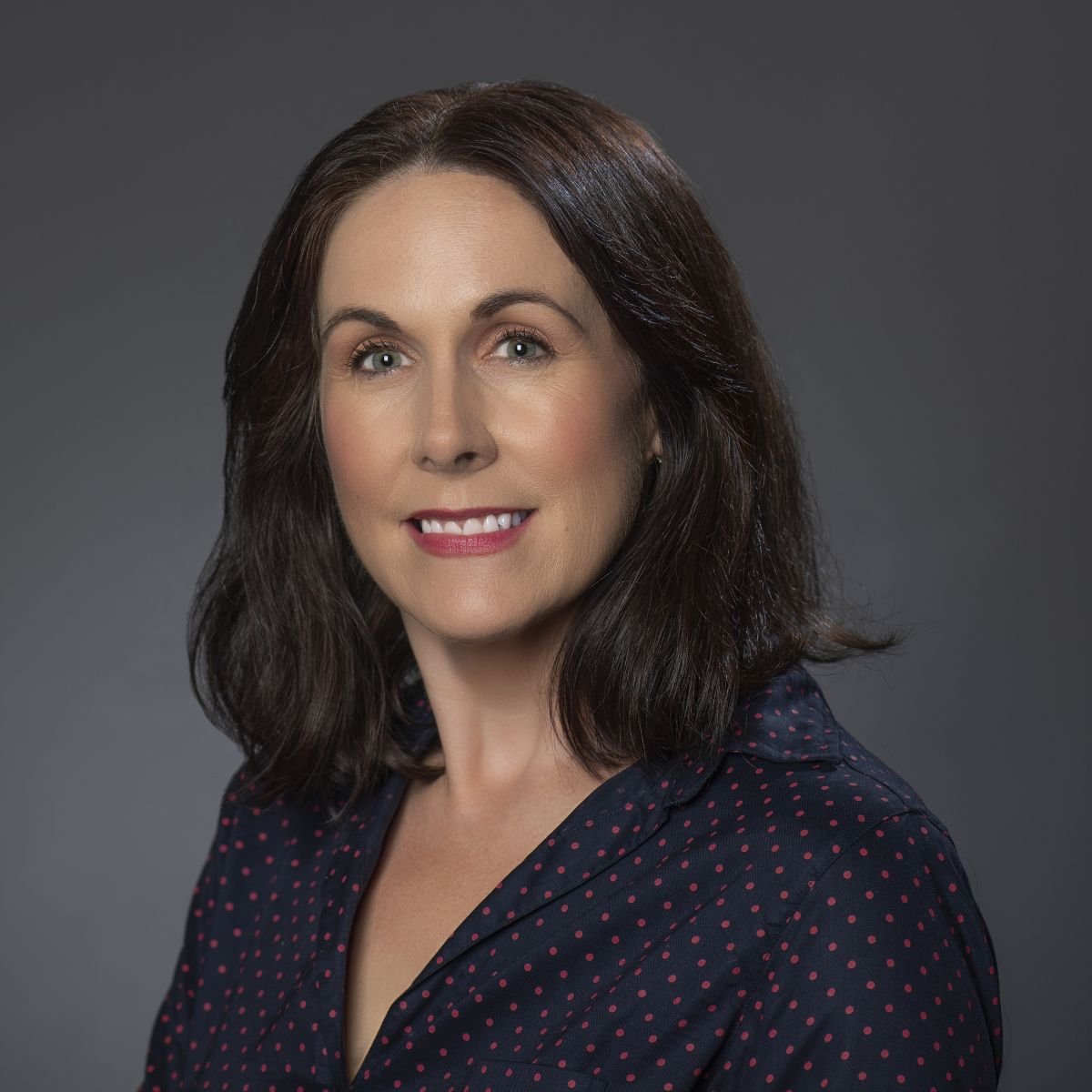 A smiling light-skinned female-presenting person with dark hair wearing a navy blue shirt against a dark gray background