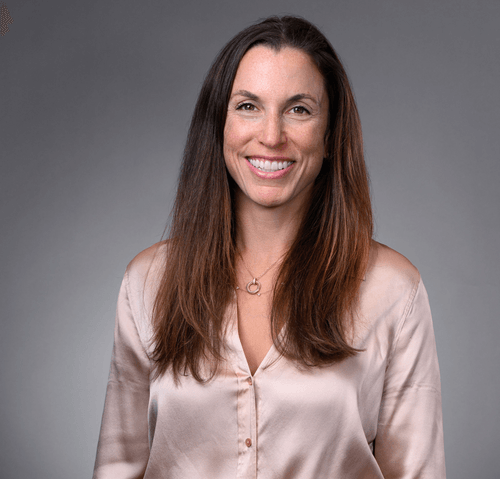 A smiling female-presenting person with long brown hair wearing a silky peach top against a gray background.