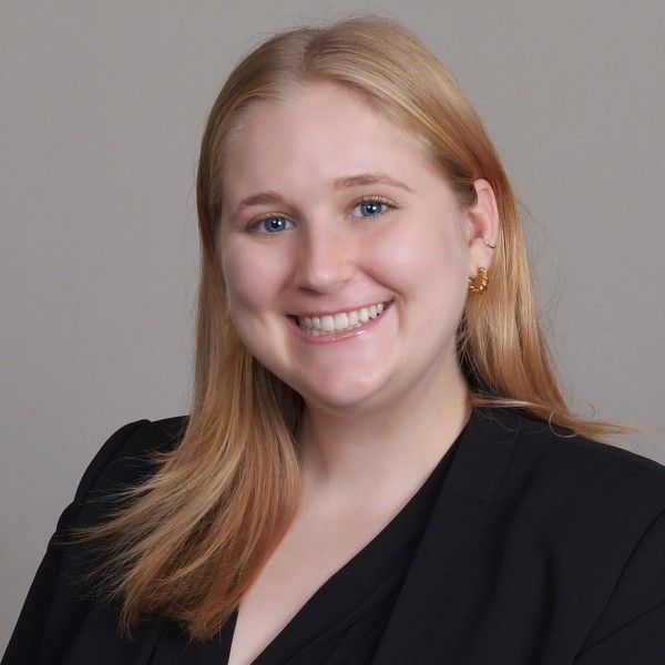 A smiling light-skinned female-presenting person with blonde hair in a black shirt against a grey background.