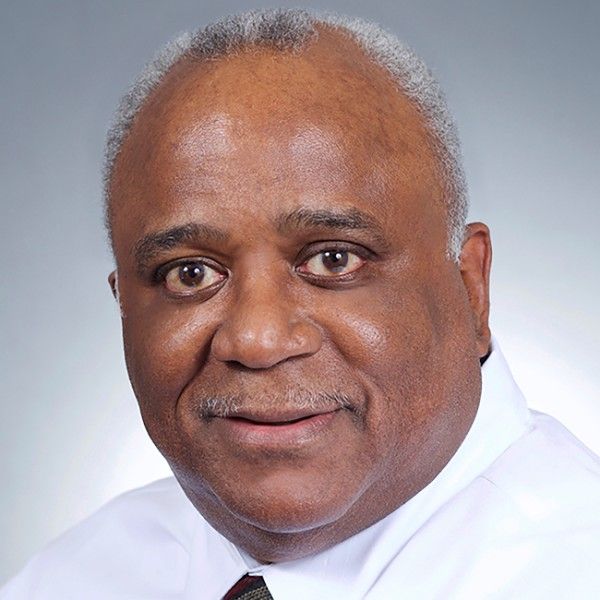 A darker-skinned, male-presenting person with gray hair wearing a button-up and tie against a light gray gradient background