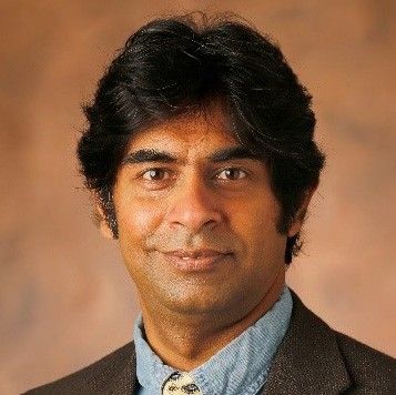 A darker-skinned, male-presenting person with black hair wearing a dark gray suit and tie against a brown gradient background