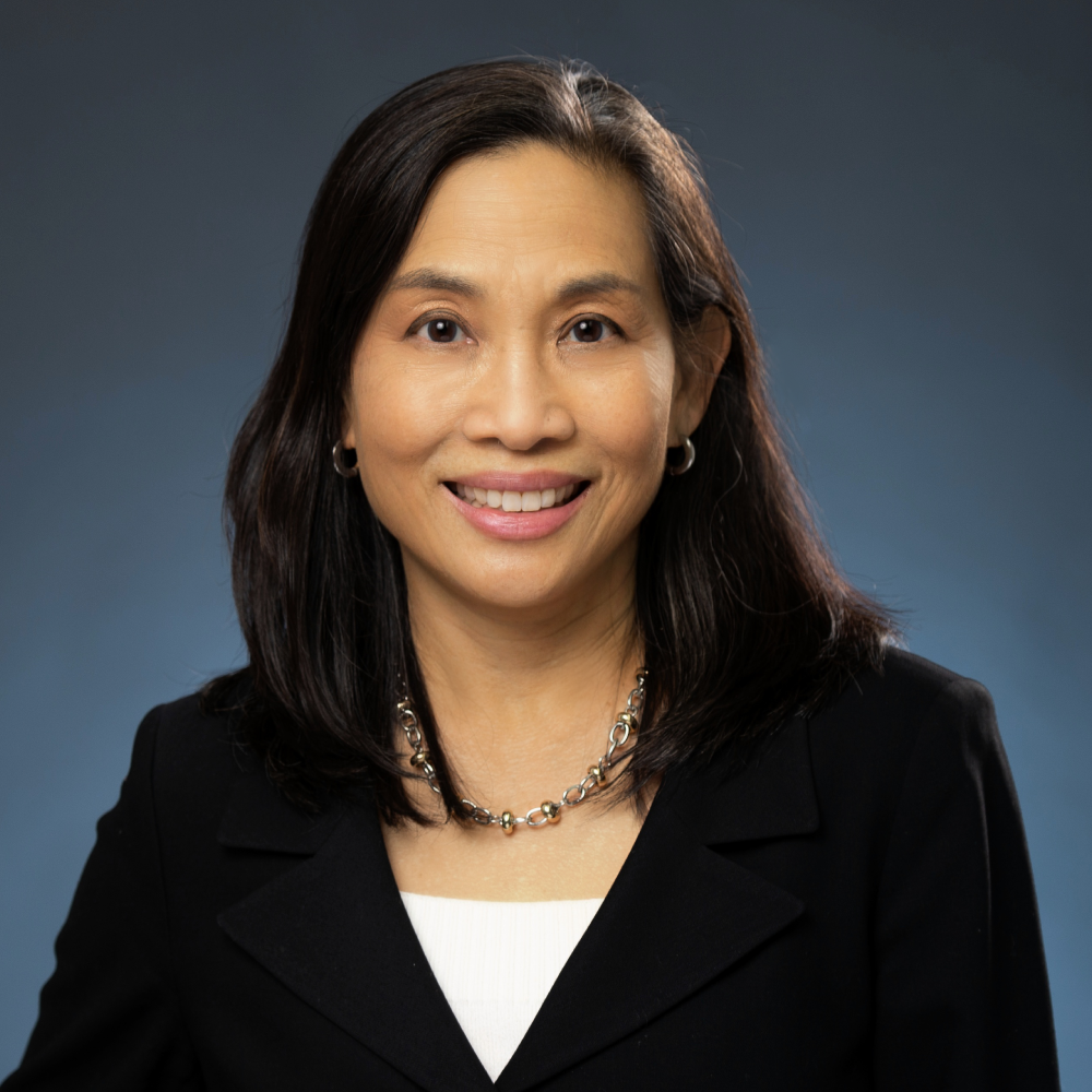 A smiling female-presenting person of Asian heritage with dark brown hair wearing a black suit jacket and a white shirt against a gray gradient background