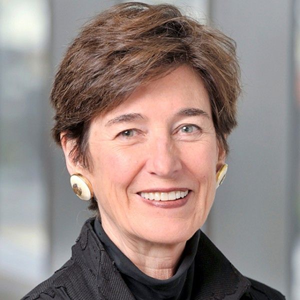 A smiling female-presenting person with short brown hair wearing a black top against a blurred background.