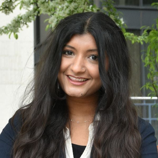 A smiling dark-skinned female-presenting person with dark hair against an outdoor backdrop.