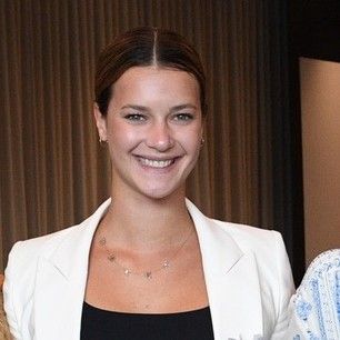 A smiling light-skinned, female-presenting individual with dark brown hair wearing a white suit jacket over a black shirt against a brown, textured background