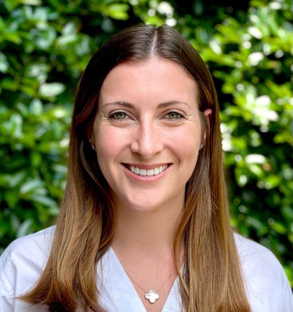 A smiling caucasian female-presenting person with brown hair wearing a white shirt against a green foliage background