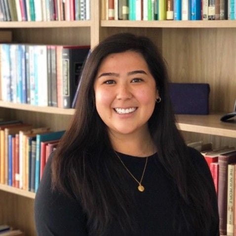 A smiling light-skinned female-presenting person with dark brown hair wearing a black shirt with a bookshelf in the background