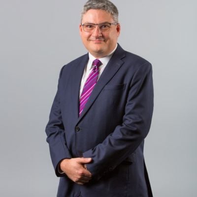 A smiling light-skinned male with gray hair wearing a dark suit and bright purple tie against a gray gradient background