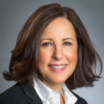 A smiling light-skinned female-presenting person with short dark brown hair wearing a button-up and suit jacket against a gray gradient background