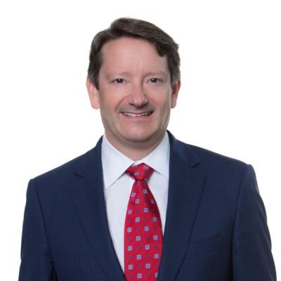 Smiling light-skinned, male-presenting individual with short dark hair wearing a dark suit on a white backdrop.